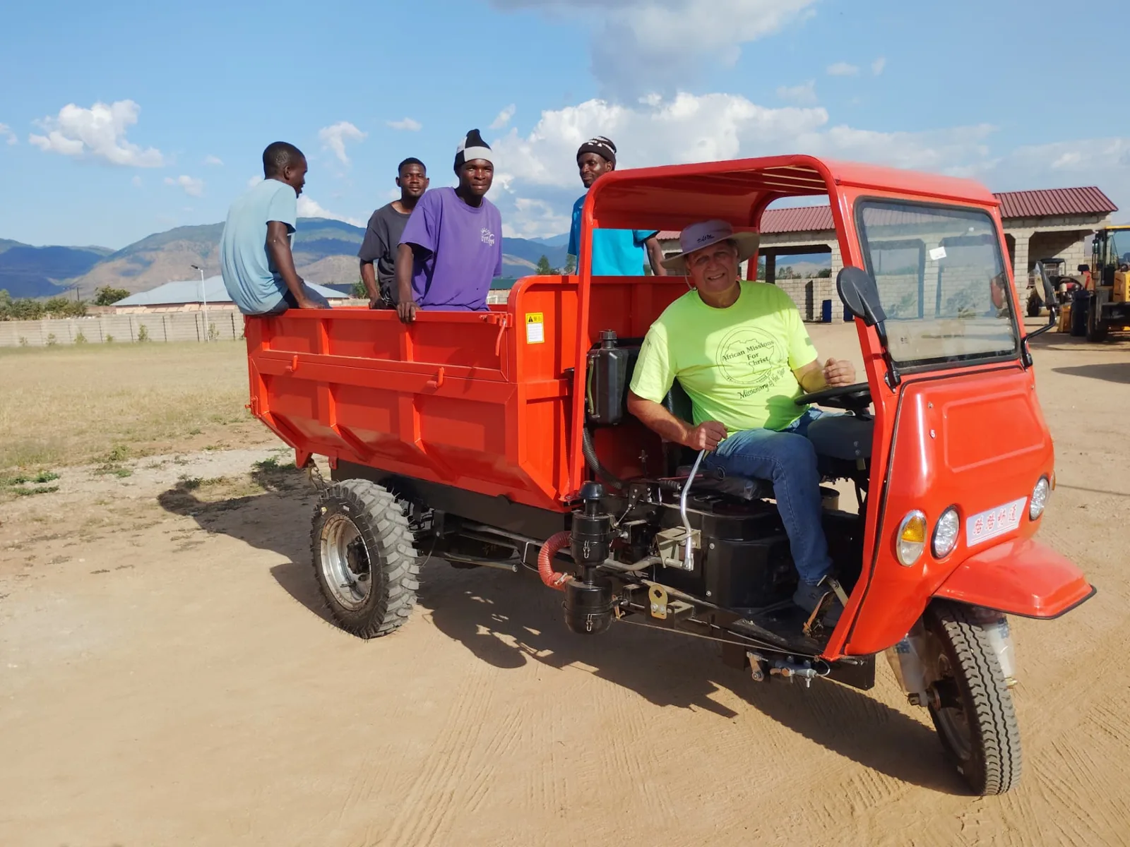 Landon Thomas driving a small orange utility dump truck with the mission crew riding in the bed on cleared ground in Africa