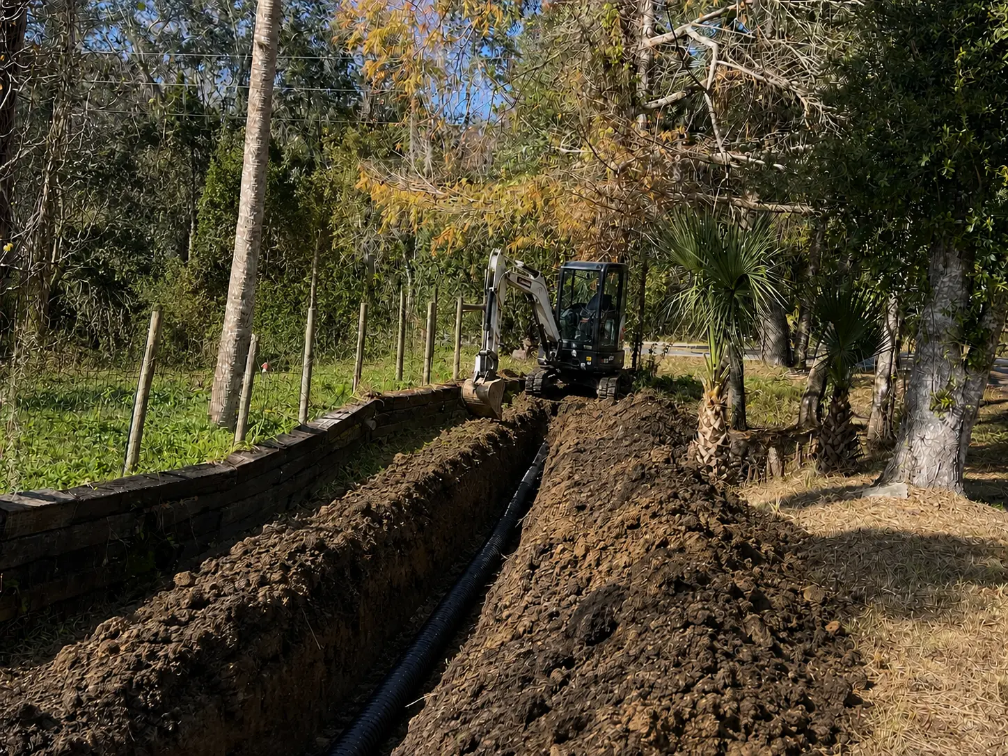 Mini excavator digging an open trench with black corrugated pipe laid in the cut during excavating work on a wooded Florida lot