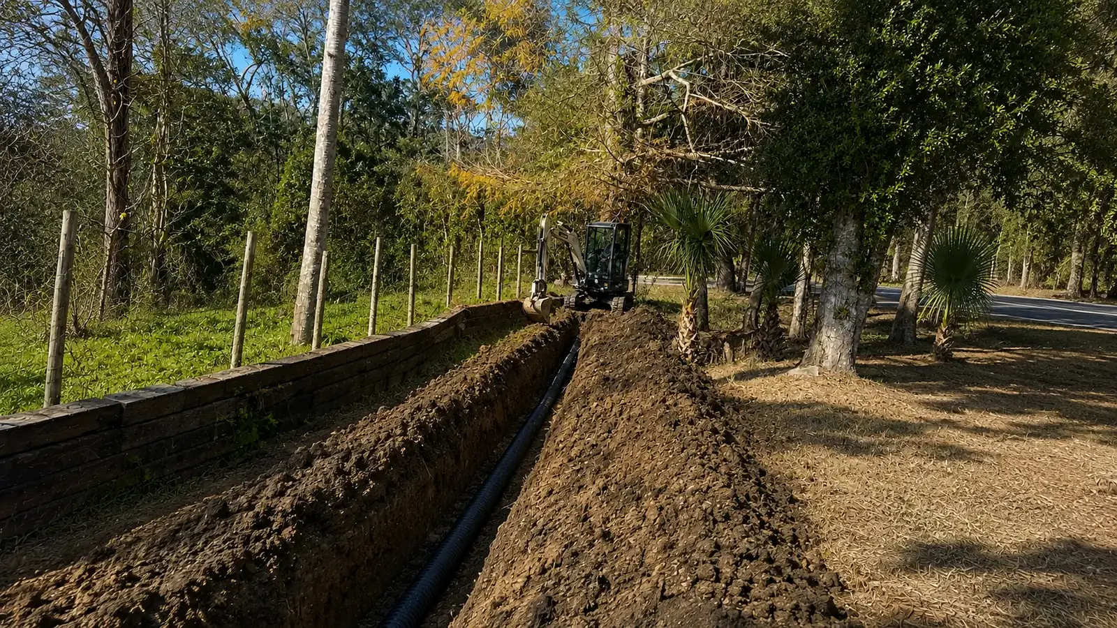 Mini excavator digs a long utility trench with black corrugated pipe laid along a fenced property line in Seminole County, Florida